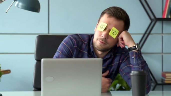 疲惫的漫画男经理假装工作时脸上贴着贴纸坐在笔记本电脑的桌子上睡觉