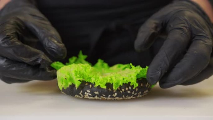 做汉堡。厨师双手戴着黑手套，将新鲜的绿色生菜沙拉放在带有酱汁的汉堡面包上。设置在黑色光泽反射台上。特