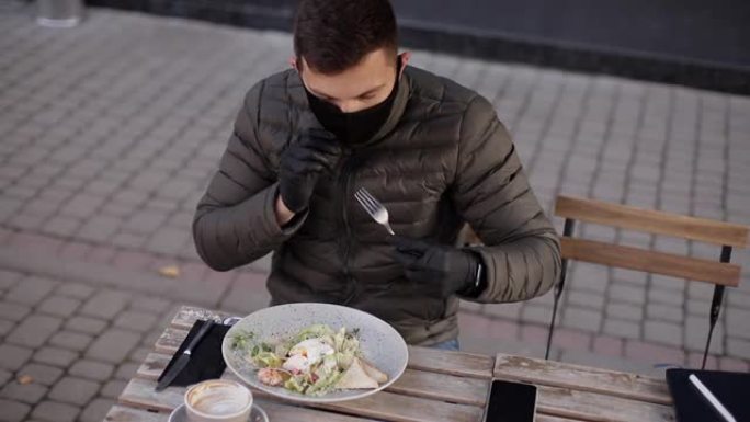人的过程为进食脱下防护面具。戴着黑手套的人拿着餐具。检疫咖啡馆概念。户外戴防护手套的食物。虾新鲜素食