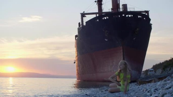 十几岁的女孩在日落时的废弃货船背景上向海水投掷石块。海岸上的旅游女孩在船上风景的傍晚日落时向水中扔石