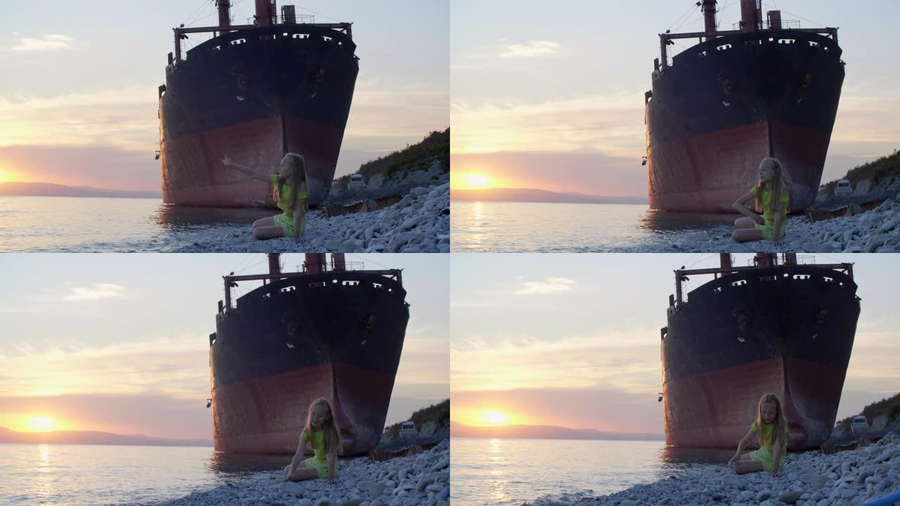 十几岁的女孩在日落时的废弃货船背景上向海水投掷石块。海岸上的旅游女孩在船上风景的傍晚日落时向水中扔石