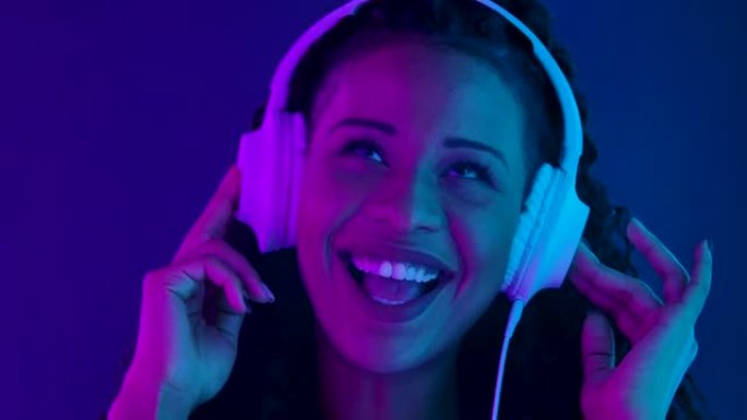 一位年轻可爱的女性非洲裔美国人的肖像看着相机，喜欢用白色大耳机听音乐。用紫色和蓝色霓虹灯照亮的特写脸