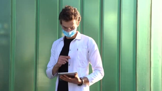 在新型冠状病毒肺炎爆发期间，白人年轻的男性医生戴着口罩，同时使用数字平板电脑对抗绿墙。高质量4k镜头