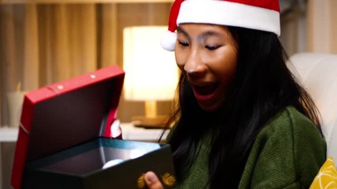 快乐的亚洲女孩穿着绿色毛衣和圣诞老人帽子坐在沙发上，晚上在家打开圣诞礼盒，节日概念。