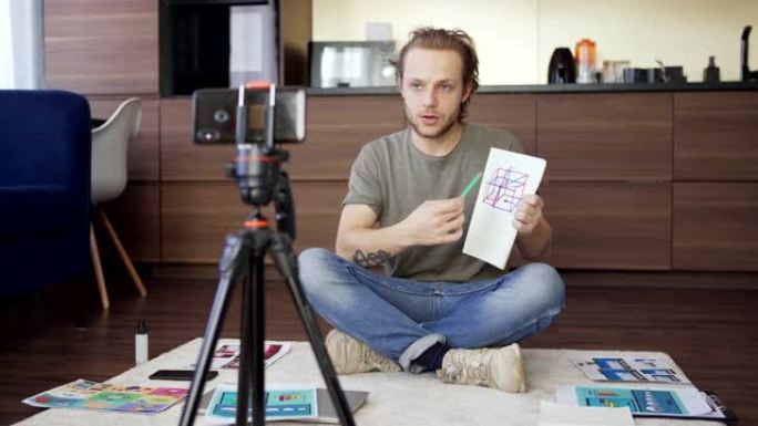 倾斜的年轻人盘腿坐在家里的地板上，与同事在智能手机上进行视频会议，并在记事本上显示他的图纸