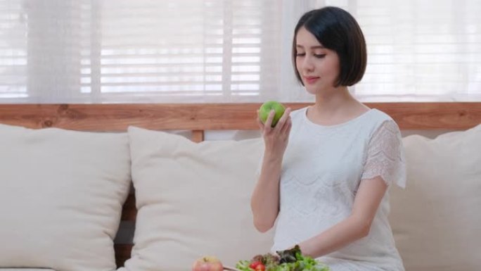 美丽的亚洲孕妇坐在客厅的沙发上，然后拿起苹果拿着也看着相机