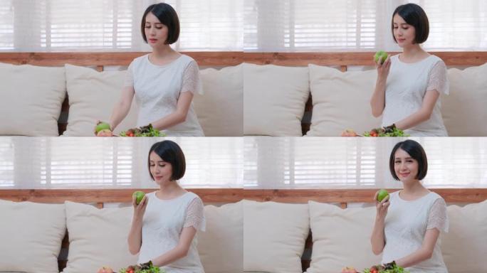 美丽的亚洲孕妇坐在客厅的沙发上，然后拿起苹果拿着也看着相机