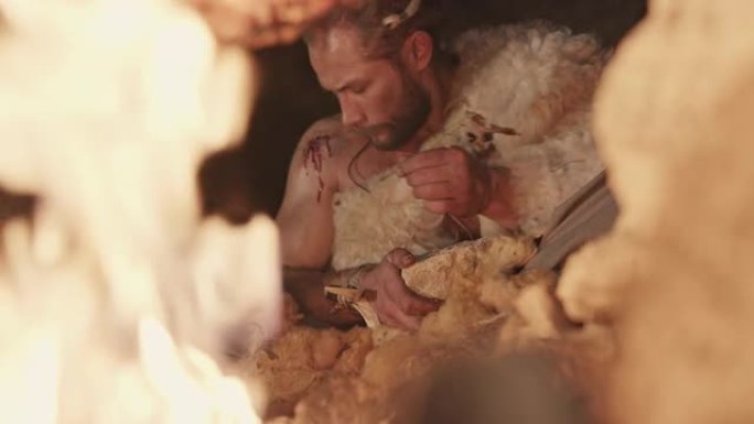 穿着原始羊皮的男人晚上坐在燃烧的篝火旁，用自己的肩膀伤口上的鲜血在石头上写字