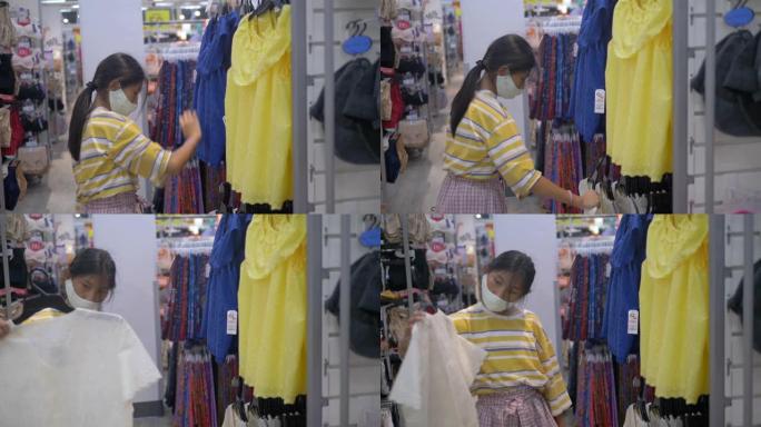 戴着面具的亚洲女孩在购物中心选择衣服，新型冠状病毒肺炎期间的新常态概念。