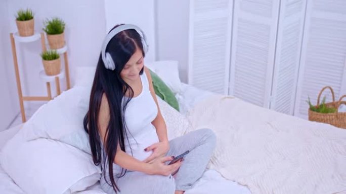 美丽的孕妇正坐在家里的沙发上，小心翼翼地抚摸着她的肚子，等待着她的第一个孩子。年轻女子戴着耳机听音乐