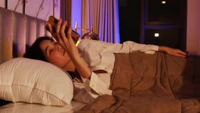 睡在家里床上的亚洲妇女醒来，禁用躺在床头柜上的手机触发的闹钟，然后转身睡在毯子里。