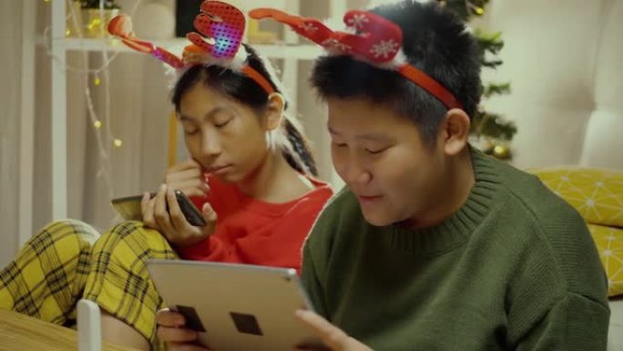 快乐的亚洲儿童穿着毛衣和发带，使用智能手机和数字平板电脑在晚上一起在家，带有圣诞节装饰背景，假期概念