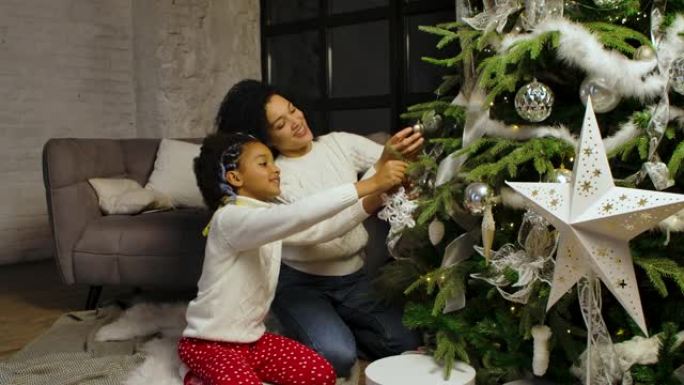快乐的非洲裔美国妈妈和小女儿的肖像用节日玩具装饰圣诞树，同时坐在阁楼风格的家庭房间的背景下。家庭之夜