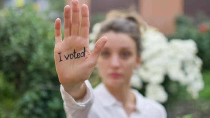 户外女性肖像的年轻成年妇女在白色衬衫显示她的手与口号“我投票了”的鲜花背景，选举日的概念