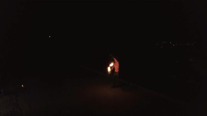 年轻的金发男性用火耍花招，晚上在黑城背景上旋转两个燃烧的火炬。旅行