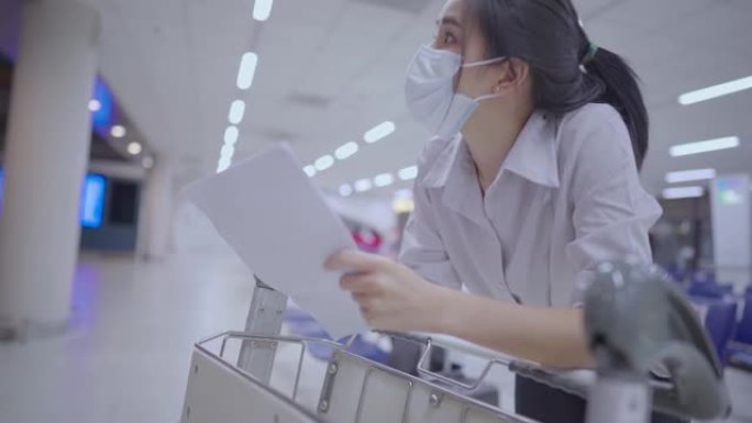 戴着医用口罩的亚洲年轻女子看着白皮书，在旅行路线文件纸上检查预订号，在空荡荡的机场航站楼，独自旅行的