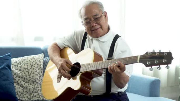 亚洲资深男子坐在沙发上弹原声吉他。快乐微笑的老人唱歌和享受吉他。在家享受退休生活。