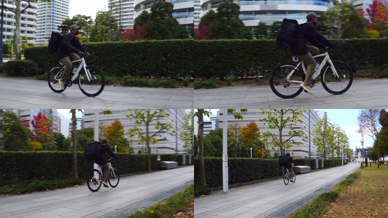 带着黑色背包和防护面具的年轻人快递员在街上骑着自行车穿过城市，运送食物。平移镜头。送餐概念。