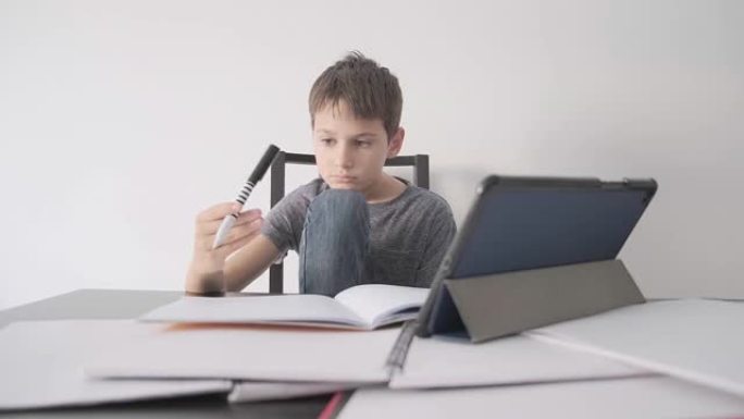 悲伤的孩子坐在桌子上，拿着平板电脑和许多书。男孩感到无聊，因为他不懂作业，他玩笔。教育，学习困难，在