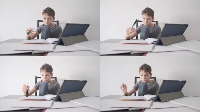 悲伤的孩子坐在桌子上，拿着平板电脑和许多书。男孩感到无聊，因为他不懂作业，他玩笔。教育，学习困难，在