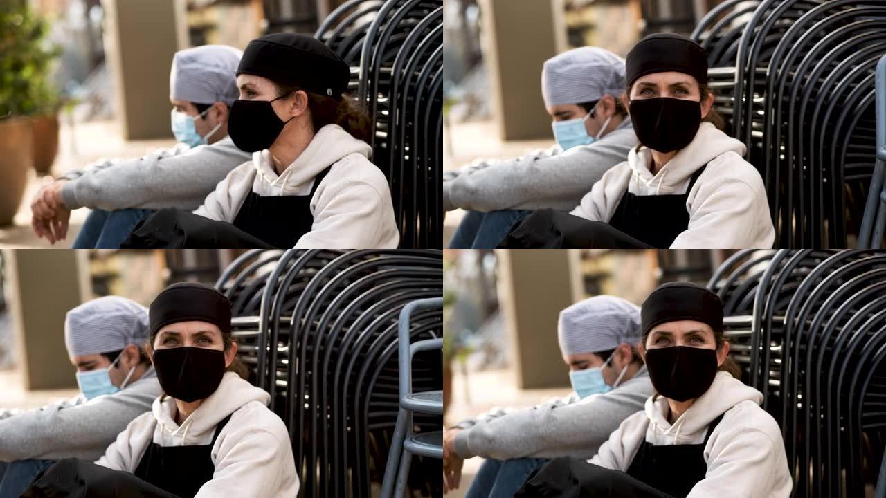 成熟的女厨师与一名男性年轻同事合影，他们戴着两个防护口罩坐在他们工作的餐厅外的地板上