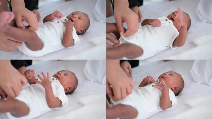 非裔美国新生婴儿母亲的双手帮助婴儿换衣服躺在白色床上，看起来放松和无辜