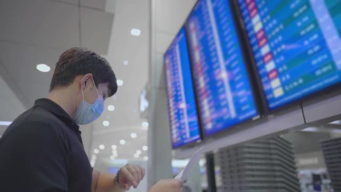 亚洲年轻男性抬头看机场航班时间表显示屏，检查旅行路线文件纸上的预订号码，在空荡荡的机场航站楼，戴口罩