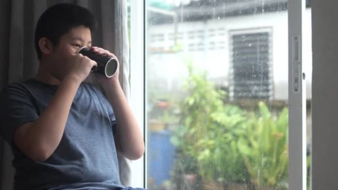 亚洲男孩在家里下雨时喝热饮料，看着窗外，生活方式理念。