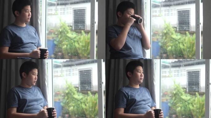 亚洲男孩在家里下雨时喝热饮料，看着窗外，生活方式理念。