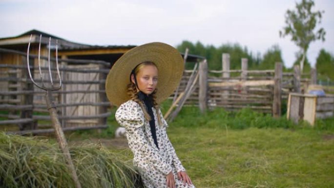 乡村的乡村女孩穿着复古连衣裙和帽子，以干草堆和干草叉为背景。乡村风景中的海瑞克帽子上的浪漫女孩肖像。