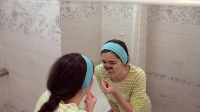 女孩从黑点上摘下黑色化妆品面膜，看着镜子里的自己，站在浴室里，护肤
