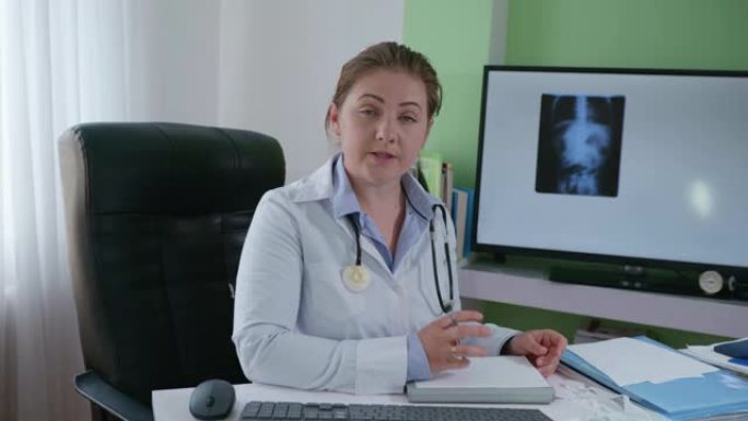 在现代医学中，一名身穿白大褂的医生带着语音内窥镜，通过带有x光图像的显示器与在线患者进行视频交流