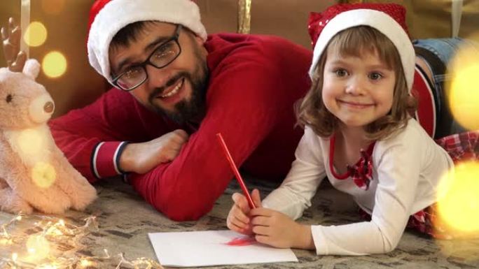 年轻父亲和戴着红色圣诞老人帽子的小女孩的肖像给圣诞老人写信给盒子的圣诞节背景。幸福的家庭庆祝圣诞节和