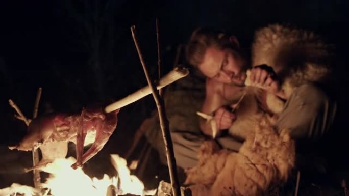 原始的狩猎采集者晚上在篝火上煮肉，一只小动物尸体在火焰上烤，他坐在那里用手工野生穴居人磨工具