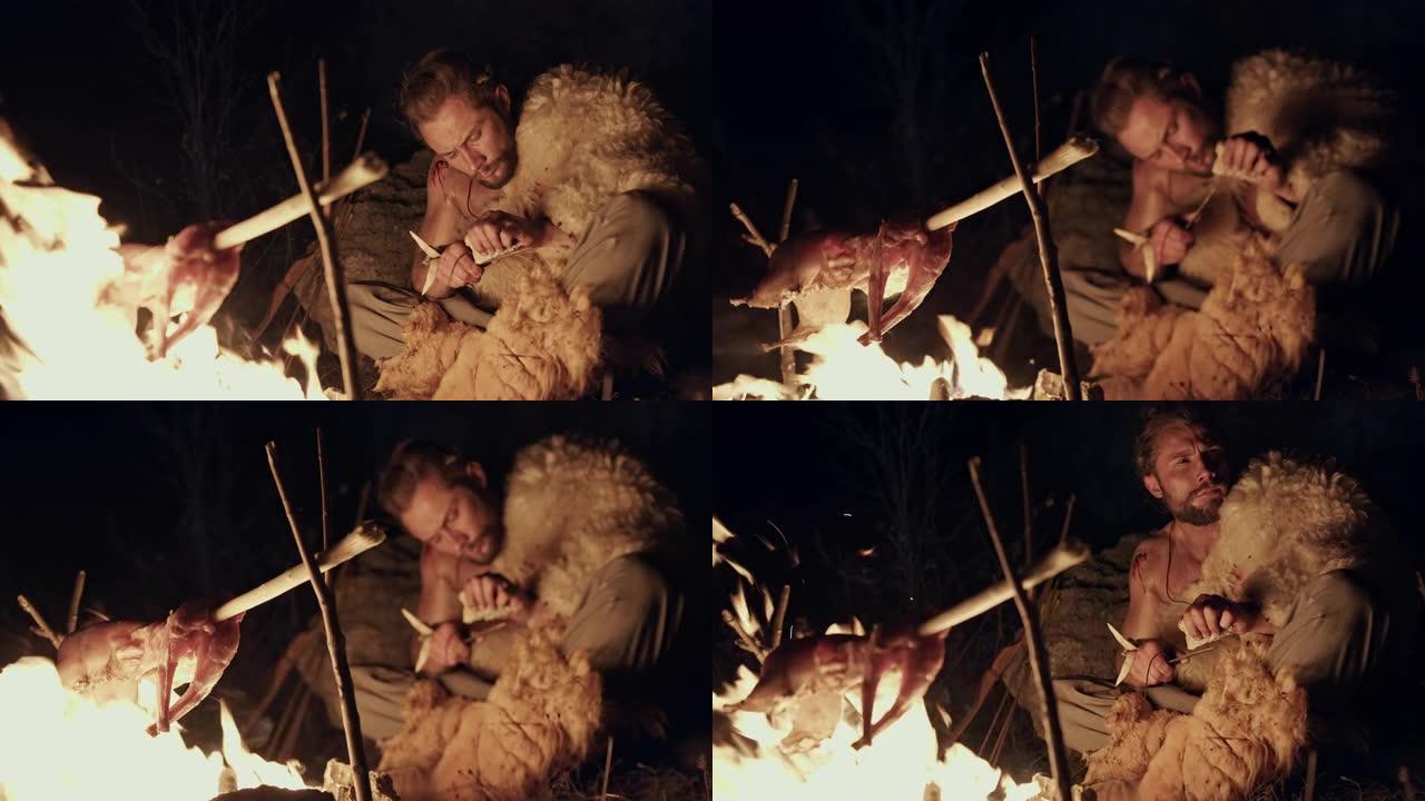 原始的狩猎采集者晚上在篝火上煮肉，一只小动物尸体在火焰上烤，他坐在那里用手工野生穴居人磨工具