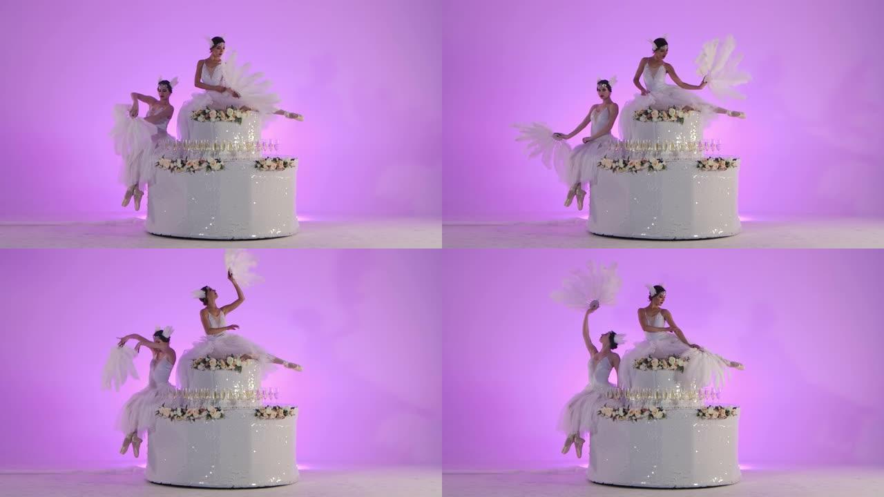 穿着白色天鹅服装的精致女性芭蕾舞演员在装饰有花朵的蛋糕上摆姿势。两名年轻女子在工作室的粉红色背景下优