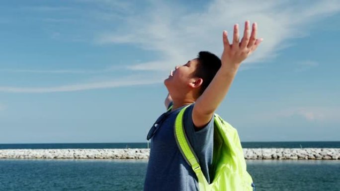 快乐的亚洲背包客男孩在天空和海洋海岸线上举手，慢动作。