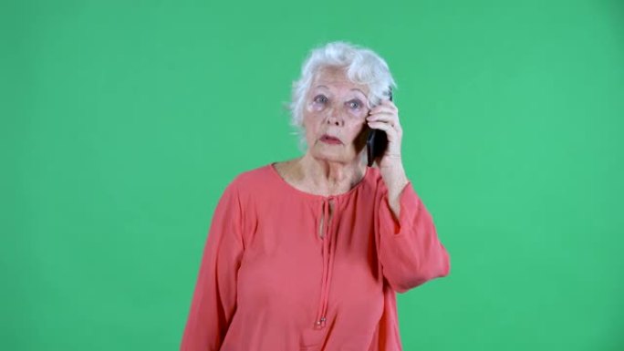 肖像老年妇女看着相机，用智能手机聊天。工作室绿色屏幕上穿着红色上衣的白发祖母。特写