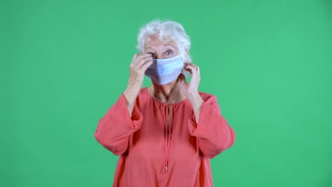 肖像老年妇女看着相机，戴着医用口罩。工作室绿色屏幕上穿着红色上衣的白发祖母。特写