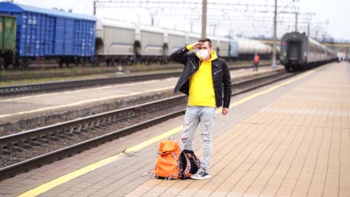 戴着医用口罩的男子站在站台上，等待火车。冠状病毒大流行期间，戴着背包的防护面罩的男性乘客在铁路平台上