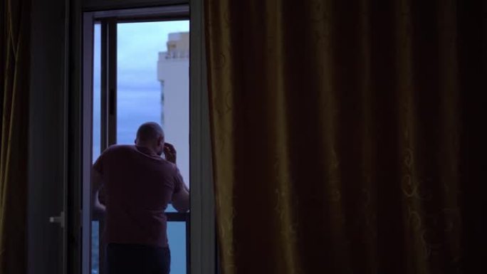 秃头男人站着，看着窗外，想着生活。它的背后是高楼和美丽的天空的一部分。从房间看。怀旧和孤独的概念