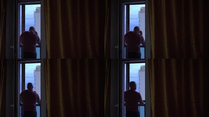 秃头男人站着，看着窗外，想着生活。它的背后是高楼和美丽的天空的一部分。从房间看。怀旧和孤独的概念