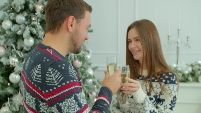 一对夫妇在发光的枞树旁拿着香槟酒杯说祝酒，祝大家一起庆祝圣诞节。圣诞夜在现代装饰的房子里浪漫的约会。