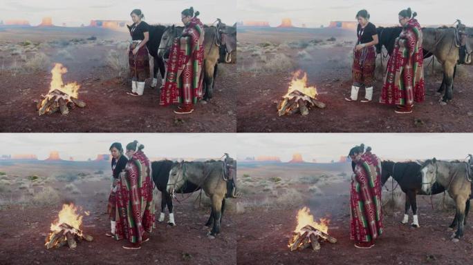 美洲原住民姐妹，一个在打鼓，包裹着传统的纳瓦霍人毯子，在他们身后的篝火马，标志性的纪念碑，日落
