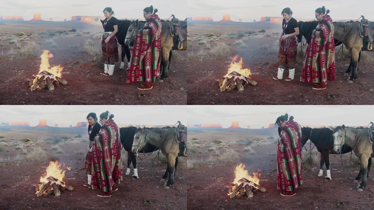 美洲原住民姐妹，一个在打鼓，包裹着传统的纳瓦霍人毯子，在他们身后的篝火马，标志性的纪念碑，日落