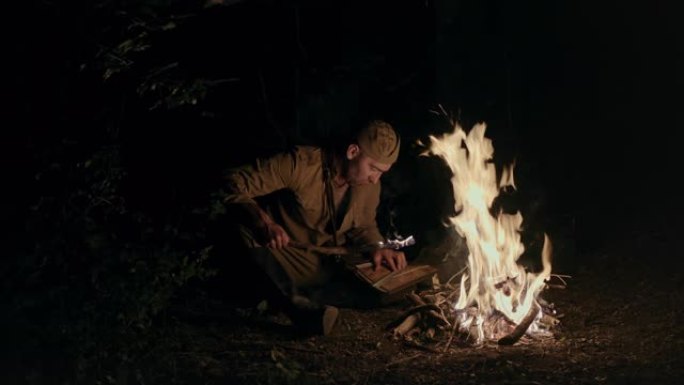 一名男性士兵在火边看地图。第二次世界大战期间，身着苏联士兵制服的成年男子坐在篝火旁，点燃火炬，查看地