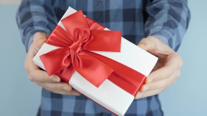 男人拿着并展示了带有红色蝴蝶结的白色礼品盒，走向相机，特写镜头。男性手中的情人节礼物。手在送礼品盒。