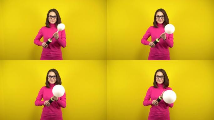 一名年轻女子用黄色背景上的泵给白色气球充气。穿着粉色高领毛衣和眼镜的女孩。
