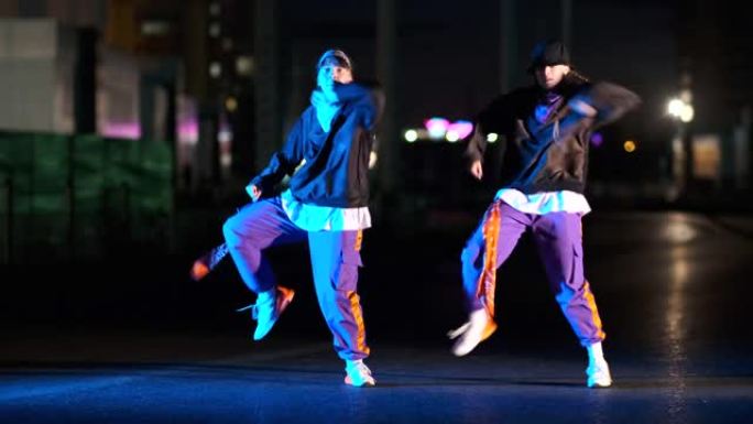 两个精力充沛的年轻女子在灯笼的灯光下，晚上在城市街道上跳着嘻哈，自由式跳舞。
