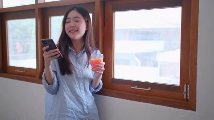 漂亮的女孩或女人用手机在玻璃窗前喝橙汁在家看起来很开心